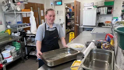 A woman with blonde hair tied in a ponytail is standing in a large kitchen holding a metal tray. 
