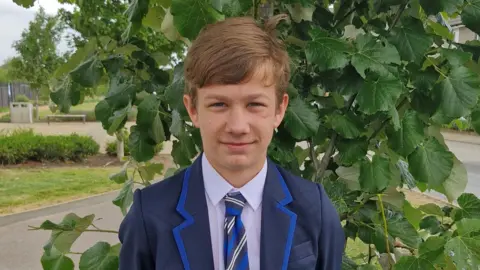 Kacper, standing outside, by a tree, smiling at the camera, wearing a blue blazer, tie and shirt. 