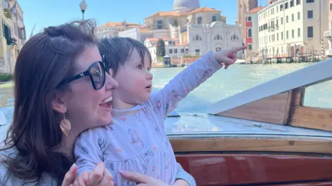 Family handout Molly aged 18 months sitting on her mother Katy's lap, pointing at something out of view. Molly wears a long sleeve pale coloured dress with foxes and flowers on. Katy has long dark straight hair and is smiling. She wears dark sun glasses and a grey coloured top.