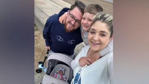 Ami Boyd Flossie is asleep in her pram while her mum, dad and brother, who are all smiling, gather together for a picture beside her. 