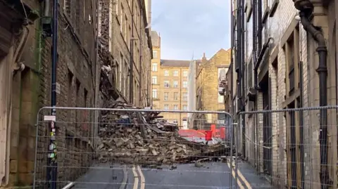 Charles Heslett/BBC A large pile of rubble in a pile blocking a road with a metal barrier fence and cordoning off the scene. There are multi-storey buildings on each side of the road. 