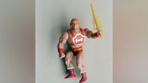 Getty Images Children's He-Man doll, 26th March 1988. (Photo by Dinello/The People/Mirrorpix/Getty Images). It shows a muscly figure wearing red boots and pants, raising a yellow sword.