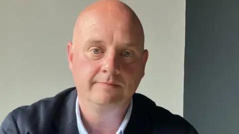 BBC A man with a bald head in a blue suit sitting with his arms in front of him