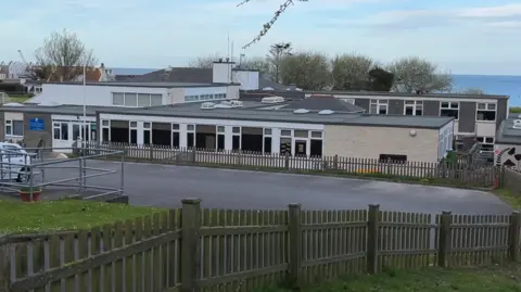 The image shows St Anne's School with a fence and car park in the foreground and the sea in the distance.