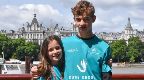 Amber Lee/PA Real Life Tom and Rosie on the day of the warrior walk, wearing bright blue T-shirts with the wording 'Cudre DHDDS'. They are smiling at the camera, stood in front of the Thames with London's old gothic architecture behind them.