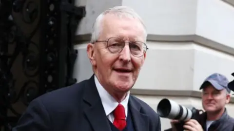 Hilary Benn - he is wearing round glasses and has short white hair. He is wearing a dark suit, dark jumper, white shirt and red tie