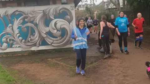 Mark Halliwell Dame Kelly Holmes, wearing a pale blue hi-vis tabard, a blue coat, black jogging bottoms and blue trainers, gives a thumbs up and smiles as she jogs through a park on a soggy winter's day in Cheltenham. Other runners can be seen taking part behind her as they run past a building with ornate swirls painted onto the side of it.