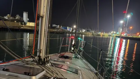 A view of the bow of a yacht, with a mast and railings at the front and the lights of a harbour in the distance during darkness.