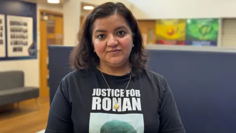 A woman wearing a “Justice for Rohan” T-shirt with a printed portrait and clip-on microphone, standing indoors in a school or community building with posters and seating in the background.
