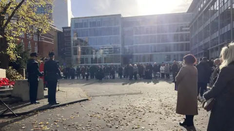 A crowd of people standing around a war memorial with wreaths of poppies laid next to it. There's a tall glass building behind the crowd and it's a sunny day.