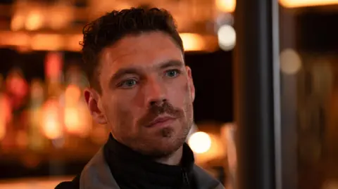 A man in a roll neck black jumper and grey jacket - inside a bar with blurry lights behind. He has facial hair and curly cropped hair.