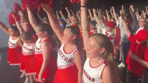 Many pupils with hands in the air during their show rehearsal of High School Musical. Some are holding red pompoms.