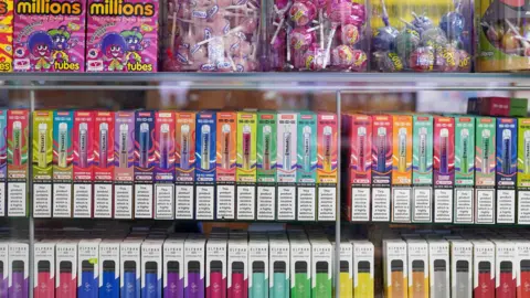 Two shelves of colourful single-use vapes in a shop. They are below sweets which have been placed on top of the glass cabinet they are in.