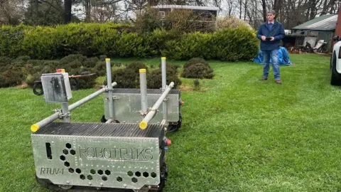 Ian Sexton is standing on grass holding a remote control. In front of him is a metal frame on wheels with the branding ROBOTRIKS.