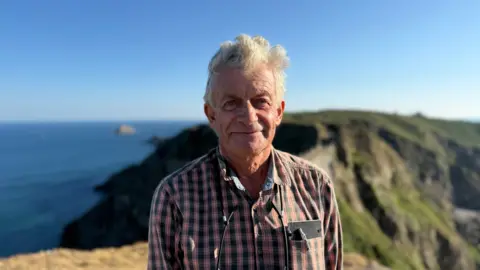 Phil Perree, a man in a black and red chequered top. He is looking at the camera and smiling. Behind him is Sark. The sky is blue. 