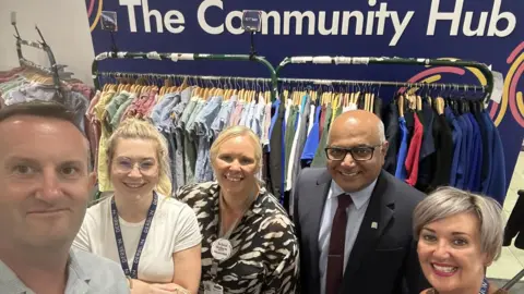 Five people smiling stood in front of a rail of school uniform. 