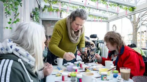 Damien Wooten A woman stands at the end of a table as she paints with a small brush. Two other women are sitting next to her. Cups and and small paint pots cover the table top.