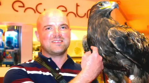 A bald man in a red, white and blue striped polo shirt with a large brown bird of prey beside him