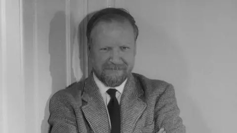A black and white picture of William Golding, who is looking at the camera with an amused expression. He has a short beard, dark hair combed over and a tweed jacket with a white shirt and a dark tie. He is leaning against a wall with a door frame visible to his left. 