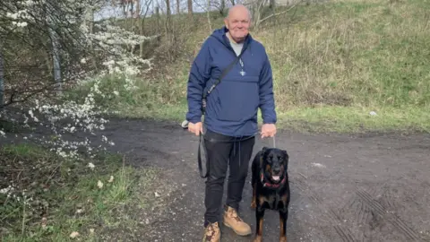 Elizabeth Baines / BBC A man wears a blue top and walks a Doberman. Trees are full of blossom to his left