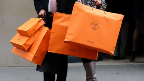 An image showing a man and a woman holding large orange Hermes branded shopping bags. Only their legs and torsos are in the frame. 