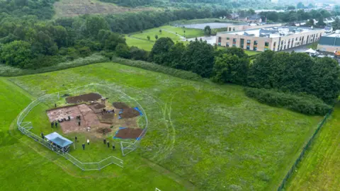Vivian Thomas Aerial shot of an excavation site at Trelai Park. A fenced off area is dug up and can be seen on the left hand corner. A group of people stand dotted around on the ground.