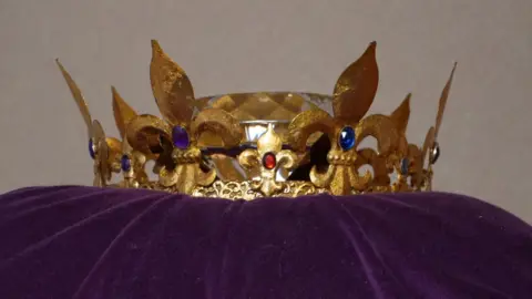 Qays Najm/BBC Golden crown with fleur-de-lys motifs around the crown's base. The upright fleur-de-lys are each decorated with a blue stone. In the middle of the crown's head lies an enormous diamond, made of paste. 