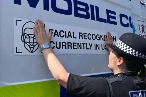 A police officer sticks a sign reading Facial Recognition Currently in Use on he side of a police van.