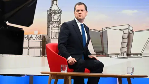 PA Media Robert Jenrick, who has short dark hair and is wearing a dark suit, white shirt and blue tie, sits in a BBC studio. There is a stylised image of the Palace of Westminster behind him and two glasses of water o a wooden table in front of him