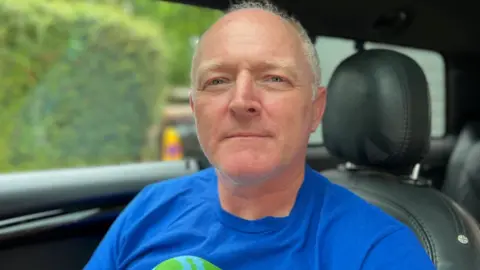 A man with grey hair and blue eyes sits in the driving seat of his car, which has a black leather-style interior. he is wearing a blue T-shirt.