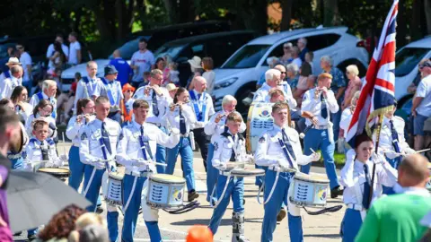 Pacemaker A large band holding drums wearing white shirts and blue trousers.