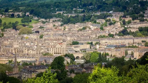 University of Bath  The city of Bath as captured from high up on a hill. You can see lots of ornate historic buildings interspersed with trees and green fields. 