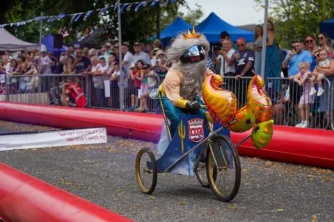 Stuart White Crowds look at a merman on a tricyle. He has a large grey beard and seahorse balloon on front of the kart.