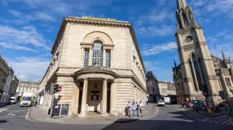 Bath & North East Somerset Council A large three-story Georgian building with a domed and pillared entrance at the end. This end of the building is narrow, but it widens from the foreground to the background.