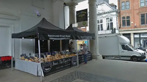 The image shows two black stalls with black awning and white writing that says "Handmade Brown Bread". Stacked loaves of bread can be seen on the stalls and  a man can be seen selling some to a woman.