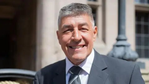 Chris Large looks at the camera, smiling, he is wearing a suit and tie. He has grey hair. He is stood infront of the county council building. 