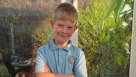 Jasper stood in front of a green bush, he has short blond hair, blue eyes and a light blue polo-shirt on. 