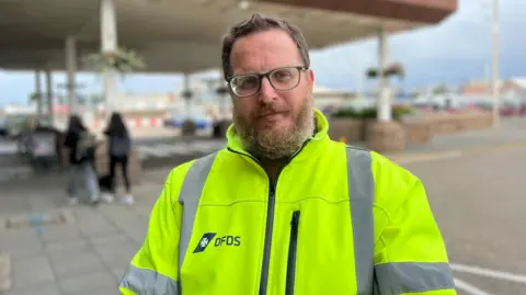 Man standing outdoors wearing a hi-vis jacket. The jacket has the DFDS logo. He has a beard and is wearing spectacles.