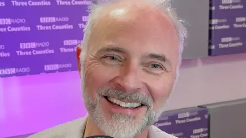Mark Bonnar smiling in a radio studio with purple BBC Three Counties Radio branding behind him.