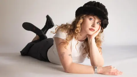 Ellur A woman in a white top black skirt and black boots lies on the floor looking a the camera. She is resting her head on one of her hands. She is wearing a dark coloured hat. She has long, curly blonde hair.