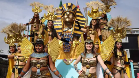 EPA Parade of ancient Egyptian dancers wearing gold bikini tops and golden wings. Some of the women also have golden headdresses with feathers attached. There is also a Tutenkhamun death mask in the background. 