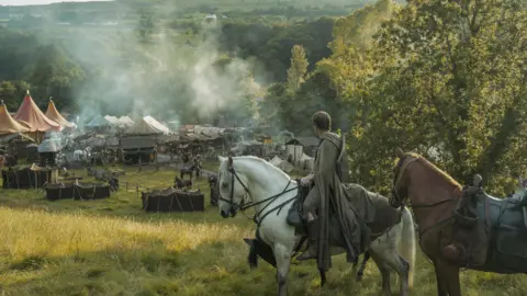 HBO Publicity shot from A Knight of the Seven Kingdoms showing Ser Duncan the Tall riding a white horse with a brown horse behind him. He has brown hair wearing a green cloak. He is looking down from a hill on towards a tourney with tents and people walking around.