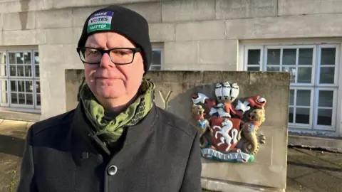 Stuart Jeffery - a man with glasses, a black coat, a black coat and a green scarf.