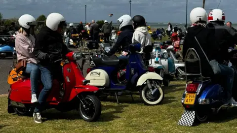 Three Vespa scooters, each with a driver and pillion passenger, on a grass verge. In the background, dozens more scooters are parked