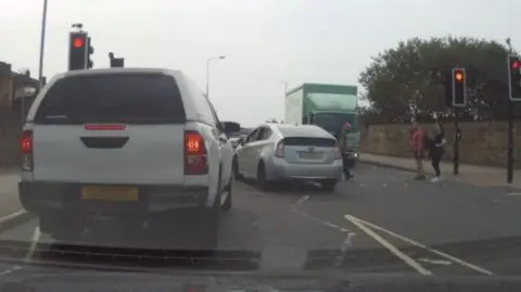 A busy road with a number of cars and one driving across a pedestrian crossing where people are trying to cross the road