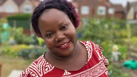 BBC Grace smiling at the camera. She is standing in an allotment in front of a housing estate surrounded by green leaves. She is wearing a red and white patterned top.