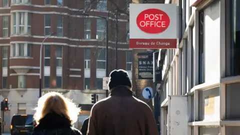 Woman with blonde hair and man wearing a woollen hat walking towards a Post Office sign 