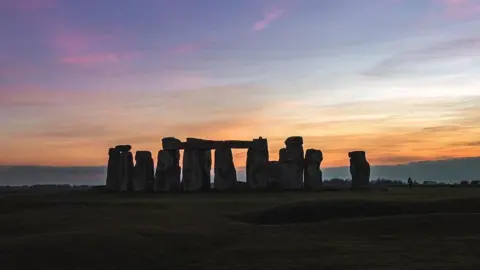 HangRoss/Weather Watchers A sunset at Stonehenge, with the stones obscured in darkness and an orange, pink and blue coloured sky in the background