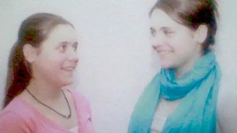 Family photo A set of twin girls with brown hair, one wearing a pink top and hair tied into a ponytail, looking at her sister wearing a blue scarf and grey top.