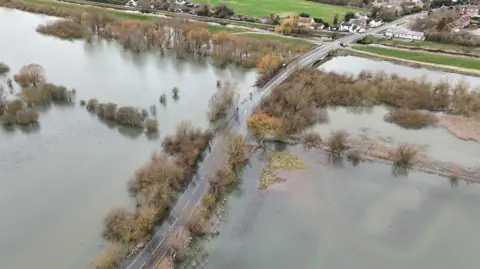 Shaun Whitmore/BBC A raised road going through a flooded landscape, with trees rising above the water. The road crosses from a village top right and drops down bottom left. 